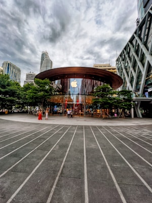 A modern glass-fronted store with a large circular wooden roof located in an urban setting. The Apple logo is prominently displayed above the entrance, which is surrounded by green trees. Skyscrapers and overcast skies are visible in the background. People are entering and exiting the store, contributing to a bustling atmosphere.