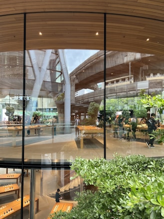 A modern, glass-fronted building with wooden interiors and greenery around it. The reflection of an overhead train track and nearby structures can be seen on the glass facade. Inside, people are gathered around large wooden tables, featuring digital devices on display.