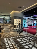 A stylish interior space with a modern grid-patterned design featuring steps and walls made from translucent glass blocks. The area is well-lit with warm artificial lighting. There is a retail display shelf filled with various products, and a plant placed in the middle of the floor near a small round table. In the background, there is a view into another section of the building, featuring a red carpet and various brand advertisements, including Givenchy.