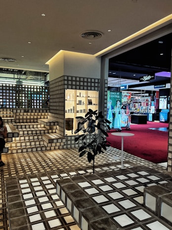 A stylish interior space with a modern grid-patterned design featuring steps and walls made from translucent glass blocks. The area is well-lit with warm artificial lighting. There is a retail display shelf filled with various products, and a plant placed in the middle of the floor near a small round table. In the background, there is a view into another section of the building, featuring a red carpet and various brand advertisements, including Givenchy.