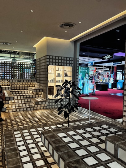 A stylish interior space with a modern grid-patterned design featuring steps and walls made from translucent glass blocks. The area is well-lit with warm artificial lighting. There is a retail display shelf filled with various products, and a plant placed in the middle of the floor near a small round table. In the background, there is a view into another section of the building, featuring a red carpet and various brand advertisements, including Givenchy.