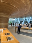 A modern retail store featuring sleek wooden furnishings and curved wooden ceiling designs. Several laptops and electronic devices are displayed on minimalist wooden tables. The store has large glass windows allowing natural light to illuminate the interior. Customers are seen browsing the products.