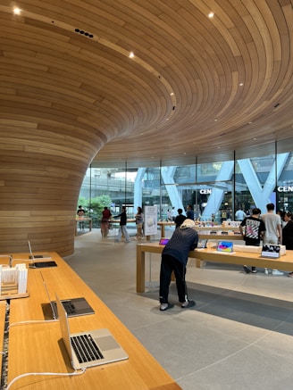 A modern retail store featuring sleek wooden furnishings and curved wooden ceiling designs. Several laptops and electronic devices are displayed on minimalist wooden tables. The store has large glass windows allowing natural light to illuminate the interior. Customers are seen browsing the products.