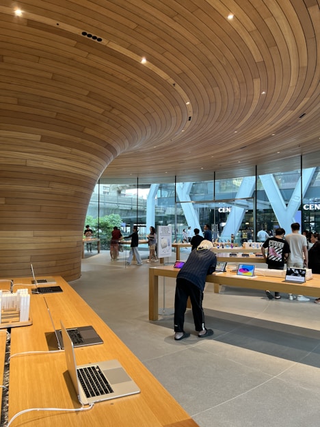 A modern retail store featuring sleek wooden furnishings and curved wooden ceiling designs. Several laptops and electronic devices are displayed on minimalist wooden tables. The store has large glass windows allowing natural light to illuminate the interior. Customers are seen browsing the products.
