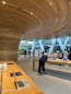 A modern retail store featuring sleek wooden furnishings and curved wooden ceiling designs. Several laptops and electronic devices are displayed on minimalist wooden tables. The store has large glass windows allowing natural light to illuminate the interior. Customers are seen browsing the products.