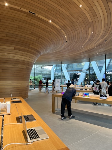 A modern retail store featuring sleek wooden furnishings and curved wooden ceiling designs. Several laptops and electronic devices are displayed on minimalist wooden tables. The store has large glass windows allowing natural light to illuminate the interior. Customers are seen browsing the products.