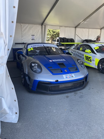 A blue and grey race car is parked inside a tent alongside another race car. Both vehicles feature sponsors' stickers and numbers on their hoods and doors. Tires are stacked nearby, and a person stands in the background wearing a hat. The scene is set in a shaded area with a bright natural light source coming from one side.
