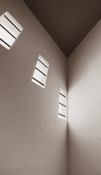 A minimalistic interior corner of a room with tall white brick walls. Three narrow vertical windows allow natural light to filter in, creating soft shadows on the walls. The ceiling features darker paneling which contrasts with the light walls. The overall aesthetic is simple and modern.