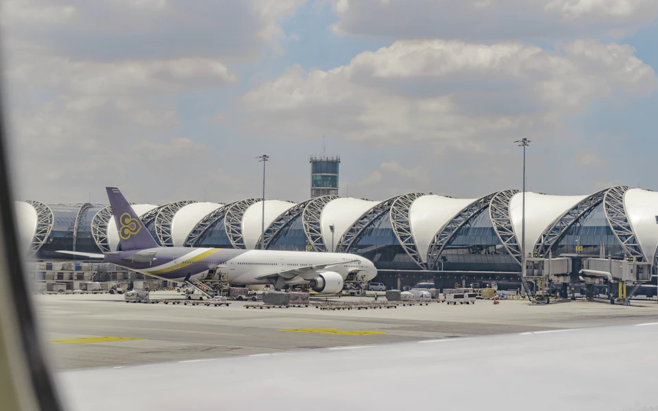 The image captures a bustling scene at an airport tarmac, with a large white airplane prominently featured on the left side of the frame. The airplane is adorned with a tail painted in hues of purple and yellow, adding a splash of color to the otherwise monochrome setting. In the background, there are several other airplanes parked or taxiing along the runway, indicating that this airport is quite busy. A few people can be seen scattered around the tarmac, likely attending to various tasks related to the aircraft's maintenance or preparation for its next flight. The sky above is a clear blue with fluffy white clouds, suggesting a bright and sunny day at the airport.