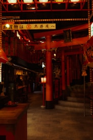 A dimly lit interior space adorned with numerous red lanterns and traditional Japanese decorations. The environment features a narrow passageway lined with red pillars and paper lanterns, giving it a cozy yet vibrant atmosphere. Several signs with Japanese characters are visible, and the overall setting appears to be a traditional market or arcade.