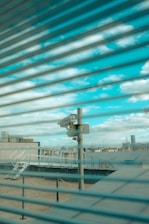 A surveillance camera is mounted on a pole overlooking a rooftop area. The scene is viewed through a set of horizontal blinds, creating a striped pattern across the image. The sky is visible in the background with scattered clouds.