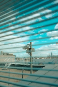 A surveillance camera is mounted on a pole overlooking a rooftop area. The scene is viewed through a set of horizontal blinds, creating a striped pattern across the image. The sky is visible in the background with scattered clouds.