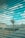 A surveillance camera is mounted on a pole overlooking a rooftop area. The scene is viewed through a set of horizontal blinds, creating a striped pattern across the image. The sky is visible in the background with scattered clouds.