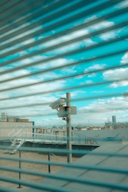 A surveillance camera is mounted on a pole overlooking a rooftop area. The scene is viewed through a set of horizontal blinds, creating a striped pattern across the image. The sky is visible in the background with scattered clouds.