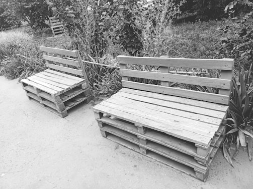 Wooden benches made from pallets are placed on a sidewalk next to shrubs and grass in what appears to be a garden or park setting. There are two benches, one larger and one smaller, constructed by stacking wooden pallets and adding a backrest made of the same material. The natural setting around the benches includes various plants and foliage.