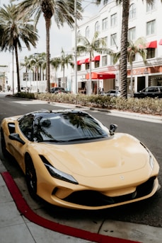 A sleek, luxury sports car with a yellow exterior is parked on a city street lined with tall palm trees. The surrounding area features high-end retail stores with red awnings and modern architecture, suggesting an upscale urban environment.