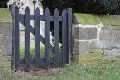 A weathered wooden fence gate with vertical slats partially open, flanked by stone walls. The gate has an oval sign that reads 'Please Close the Gate'. Lush greenery and the corner of a historical stone building are visible in the background.