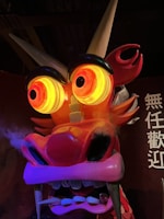 A large, colorful dragon head with large, illuminated eyes and a fierce expression. The dragon has prominent red and orange hues, with sharp teeth and smoke or mist emanating from its mouth. The horns are long and pointed, adding to the intimidating look. Chinese characters are visible in the background.