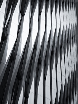 The image features a modern architectural facade with a geometric, repeating pattern of overlapping and protruding panels. The design creates a sense of depth and dynamism, with sharp angles and shadows accentuating the three-dimensional effect.