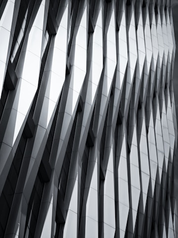 The image features a modern architectural facade with a geometric, repeating pattern of overlapping and protruding panels. The design creates a sense of depth and dynamism, with sharp angles and shadows accentuating the three-dimensional effect.