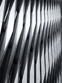 The image features a modern architectural facade with a geometric, repeating pattern of overlapping and protruding panels. The design creates a sense of depth and dynamism, with sharp angles and shadows accentuating the three-dimensional effect.