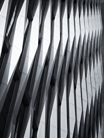 The image features a modern architectural facade with a geometric, repeating pattern of overlapping and protruding panels. The design creates a sense of depth and dynamism, with sharp angles and shadows accentuating the three-dimensional effect.