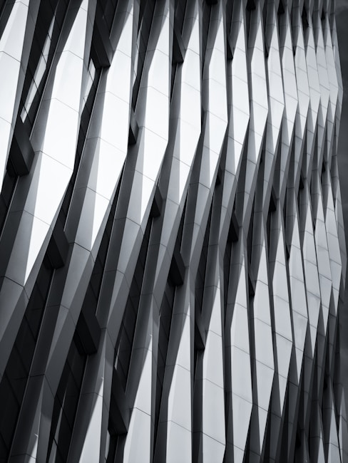 The image features a modern architectural facade with a geometric, repeating pattern of overlapping and protruding panels. The design creates a sense of depth and dynamism, with sharp angles and shadows accentuating the three-dimensional effect.