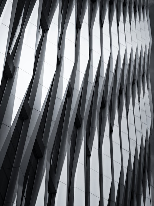 The image features a modern architectural facade with a geometric, repeating pattern of overlapping and protruding panels. The design creates a sense of depth and dynamism, with sharp angles and shadows accentuating the three-dimensional effect.