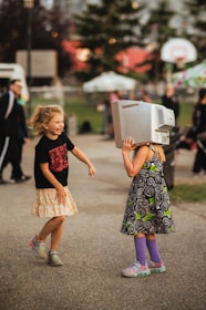Two children are playing outdoors on a paved path. One child is wearing a black t-shirt and a patterned skirt, laughing and looking at the other. The other child has a computer monitor on their head, wearing a dress with a swirl pattern, and colorful shoes and socks. The background features trees, a basketball hoop, and blurred figures, suggesting a park or community event.