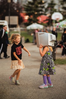 Two children are playing outdoors on a paved path. One child is wearing a black t-shirt and a patterned skirt, laughing and looking at the other. The other child has a computer monitor on their head, wearing a dress with a swirl pattern, and colorful shoes and socks. The background features trees, a basketball hoop, and blurred figures, suggesting a park or community event.