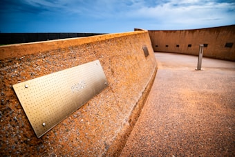 A curved, rust-colored wall with a metallic plaque featuring the engraved word 'PEACE.' The ground is textured in a similar color, and the sky above is overcast with shades of blue and gray. A series of smaller plaques and metal posts are distributed along the wall.