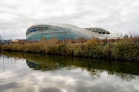 A modern, architectural stadium with a sleek, curved design is situated by a body of water. The structure has a metallic and glass exterior, reflecting the cloudy sky above. Dense shrubbery lines the water's edge, adding splashes of earthy tones to the scene.