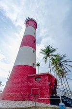 A tall, striped lighthouse with red and white bands stands prominently against a partly cloudy sky, surrounded by tall palm trees. The structure has a modern, well-maintained appearance with a paved path leading up to it.