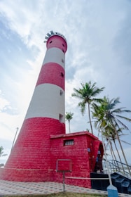 A tall, striped lighthouse with red and white bands stands prominently against a partly cloudy sky, surrounded by tall palm trees. The structure has a modern, well-maintained appearance with a paved path leading up to it.