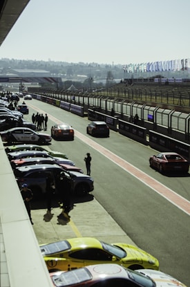 A racetrack scene featuring several parked cars along a pit lane, with people standing around and a few vehicles driving on the track. Numerous flags line the fence along the track, and the background shows grandstands and a distant view of the city skyline.