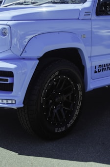 A close-up of the front side of a light blue SUV with custom black wheels featuring Cooper Discoverer AT3 tires. The vehicle has a modified body kit with visible branding on the side. The lighting highlights the smooth surface and angular design of the car's exterior.