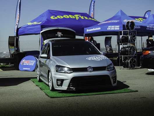 A silver Volkswagen car is prominently displayed in front of a blue tent with a Goodyear logo. The setup appears to be part of an outdoor automotive event, with spare tires neatly stacked to the side. Promotional banners and additional signage for Tyremart are visible around the area, and there are other cars in the background.
