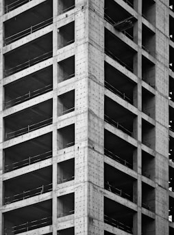 A partially constructed high-rise building with visible concrete columns and beams forming a grid pattern. The structure is incomplete, with open spaces where windows and walls are yet to be installed.