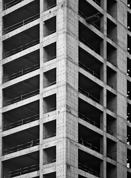 A partially constructed high-rise building with visible concrete columns and beams forming a grid pattern. The structure is incomplete, with open spaces where windows and walls are yet to be installed.