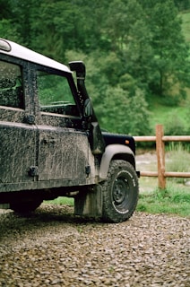 A rugged, mud-splattered off-road vehicle is parked on a gravel path in a lush, green countryside setting. The vehicle's black and white exterior suggests it has been used for outdoor adventures. A wooden fence runs parallel to the vehicle and adds to the rural atmosphere. The background features dense green foliage and trees, indicating an outdoor natural environment.