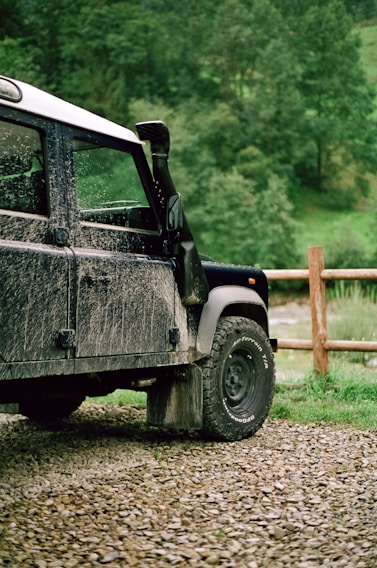 A rugged, mud-splattered off-road vehicle is parked on a gravel path in a lush, green countryside setting. The vehicle's black and white exterior suggests it has been used for outdoor adventures. A wooden fence runs parallel to the vehicle and adds to the rural atmosphere. The background features dense green foliage and trees, indicating an outdoor natural environment.