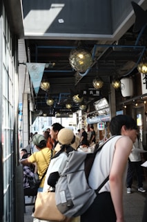 A busy street market scene with people walking and shopping under decorative lighting. Various stalls are visible along the right, with vendors attending to customers. Some people are carrying backpacks, and there's a woman wearing a straw hat. The atmosphere appears lively and bustling.