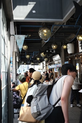 A busy street market scene with people walking and shopping under decorative lighting. Various stalls are visible along the right, with vendors attending to customers. Some people are carrying backpacks, and there's a woman wearing a straw hat. The atmosphere appears lively and bustling.