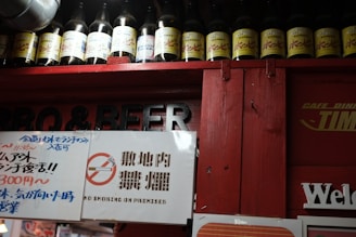 A row of bottles with yellow labels is arranged on a shelf above a red structure. Signs with various texts, including a no smoking sign, are displayed below in both Japanese and English.
