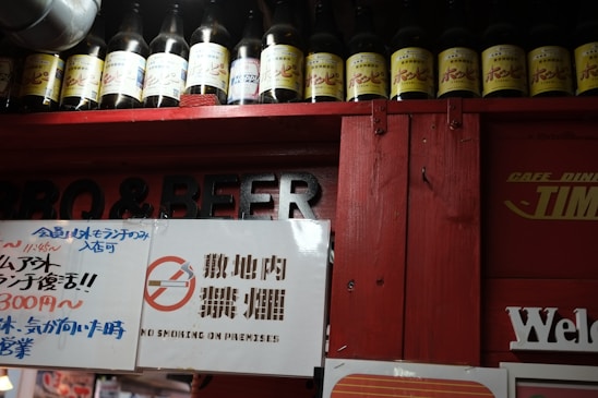 A row of bottles with yellow labels is arranged on a shelf above a red structure. Signs with various texts, including a no smoking sign, are displayed below in both Japanese and English.