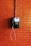 A vintage public telephone mounted on a brick wall. The phone is black with a coiled cord and a numeric keypad. A pipe runs vertically above the phone.