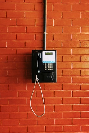 A vintage public telephone mounted on a brick wall. The phone is black with a coiled cord and a numeric keypad. A pipe runs vertically above the phone.