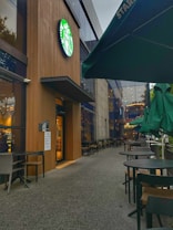 The setting is an outdoor seating area of a café with a modern exterior. The iconic green logo of a well-known coffee chain is prominently displayed above the entrance. Several tables and chairs are arranged on the patio, and green umbrellas are closed, suggesting an outdoor dining option.