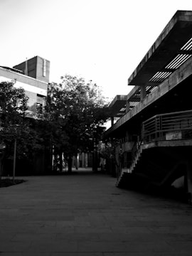 A black and white photograph featuring an urban environment with modern architecture. The scene includes concrete buildings and structures with staircases and railings, alongside trees lining an empty pathway. Shadows and lighting contribute to a moody atmosphere.