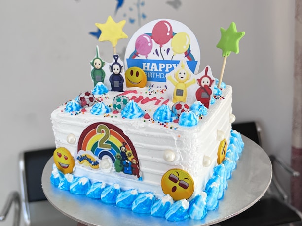 A colorful birthday cake adorned with various decorations, including cartoon character toppers, emoji faces, and a rainbow with the number two. The cake features bright blue frosting accents, star-shaped decorations, and a circular 'Happy Birthday' sign with balloons. The overall theme appears to be playful and vibrant.